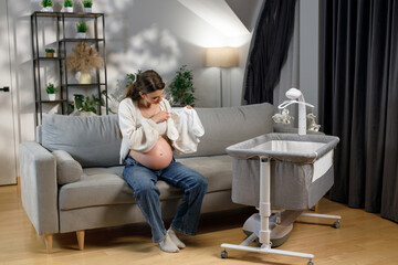 Expecting mother sits on sofa holding baby clothes near bedside bassinet preparing for newborn arrival