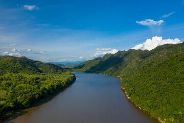 Obraz premium The Mekong river curves between densely forested hills under a vivid blue sky with scattered clouds. Sunlight highlights the lush vegetation along the riverbanks, creating a serene and expansive