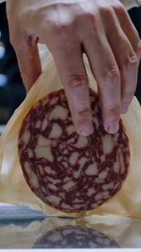 Hands arranging round slices of artisanal salami on a beige paper lining inside clear vacuum packaging, preparing food for retail or storage, demonstrating preparation
