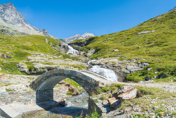 Ancient bridge made of stone on the path leading to "San Grato" lake, Valgrisenche, Aosta Valley, Italy. High mountains peaks on the background. Green meadow on the right.