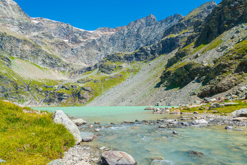 The waters of "San Grato" lake, Aosta Valley, Italy, with water flowing in the foreground. High peaks on the background with blue sky.