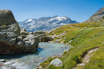 High mountain water stream in Aosta Valley, Italy. Green meadow with hiking path on the right. High peaks with glaciers and snow on the background, with blue sky.