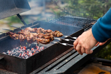 Man cooking outdoors, holding skewers with pieces of meat above glowing coals on grill, preparing food on backyard of house in the mountains