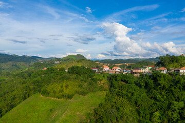 Fototapeta premium A cluster of colorful houses sits along a verdant hillside in Phou Khoun, Laos, surrounded by rolling green hills and dense vegetation under a wide blue sky with dramatic clouds. The scene highlights