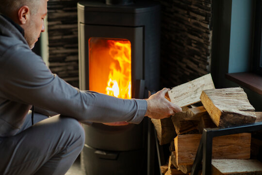 Man stacking firewood near wood stove that burning brightly in room, heating house in cold season