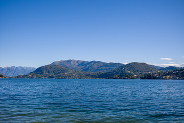 Fototapeta premium Wide view across Lake Orta in northern Italy, featuring deep blue water and gentle mountain slopes beneath a cloudless sky. The distant shoreline is dotted with small villages and lush green hills