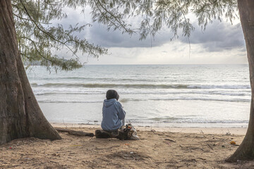 Fototapeta premium Lone person sits on the sandy beach framed by trees using the phone and watching the calm sea and distant horizon. Leisure, sea watching