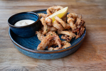 Crispy squid calamari, deep fried and dusted with salt and pepper alongside a bowl of herb aioli. The food is in a blue dish and on a wooden table.