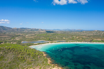 Fototapeta premium Aerial view of Cala Brandinchi in Sardinia, featuring a shallow turquoise lagoon, white sandy beach, and dense pine forest with rolling hills and distant mountains under a clear blue sky.