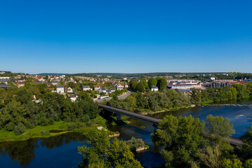 Wide aerial perspective of Imphy in central France, featuring a bridge crossing the Loire river, lush green trees, and a mix of residential and industrial buildings under a vivid blue summer sky. The