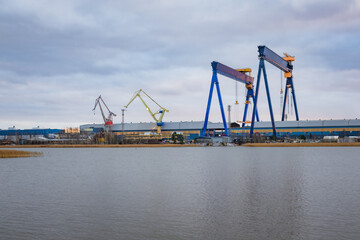 Obraz premium Photo of a large ship being constructed at a shipyard near the sea, captured from a distant vantage point. Shows cranes, scaffolding, massive vessel structure, and industrial maritime environment.