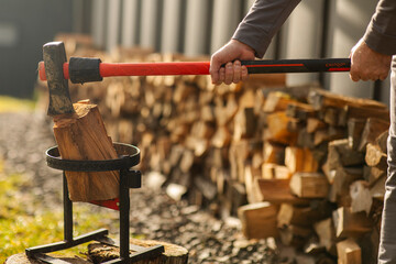Man preparing to split piece of wood using large axe on backyard with stacks of firewood on background, closeup shot