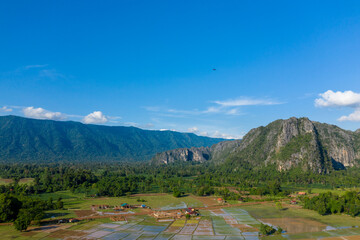 Fototapeta premium Aerial view of dramatic limestone cliffs rising above a mosaic of flooded rice fields and scattered rural houses, set against lush forest and distant mountains under a vivid blue sky. The landscape