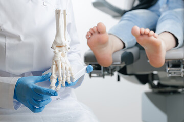 Podiatrist doctor showing foot skeleton model on background injured leg of patient in traumatology clinic white light