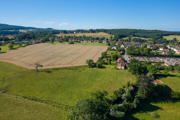Naklejka premium Aerial view of a golden harvested field meeting the edge of Saint-Honore-les-Bains village, surrounded by lush greenery, scattered houses, and distant forested hills under a bright blue sky.