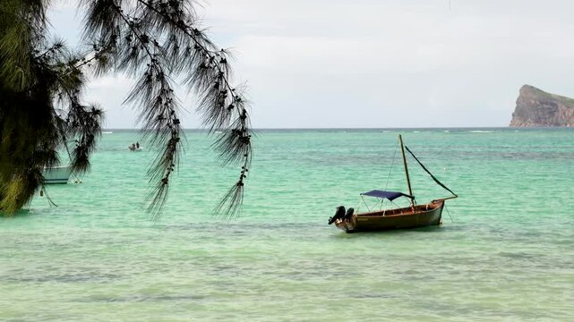 Traditional wooden boat anchored in the turquoise lagoon of Mauritius, framed by green pine branches with the iconic Coin de Mire rock in the background scenery