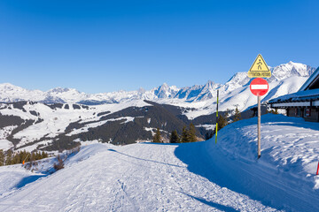 Bright winter scene featuring a snow-covered ski trail overlooking forested hills and dramatic alpine peaks under a vivid blue sky near Megeve in the French Alps. Warning signs add a touch of color