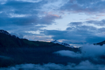 Naklejka premium Dramatic blue clouds drift above the snow-dusted summit of Mont Joly, with mist swirling over dark pine forests in the tranquil evening light.