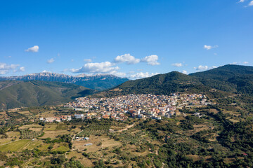 Naklejka premium Wide aerial view of Orgosolo nestled among green hills and rugged mountains, under a bright blue sky with scattered clouds in Sardinia, Italy.