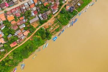 Aerial view of vibrant longboats moored along the muddy bank of the Nam Ou River, with rustic village rooftops and lush greenery in Nong Khiaw, northern Laos. The image highlights the vivid colors