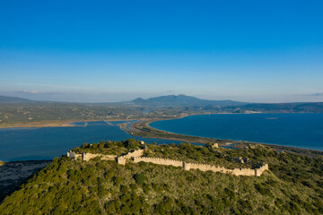 Drone view of ancient stone castle ruins stretching along a green, forested hilltop overlooking a sweeping blue bay near Navarino, Greece, under a bright, cloudless sky.
