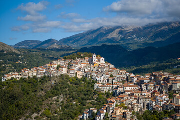 Fototapeta premium Dense arrangement of traditional stone houses with terracotta roofs in Rivello, Italy, set on a sunlit hillside with lush greenery and rugged mountains under a partly cloudy sky.