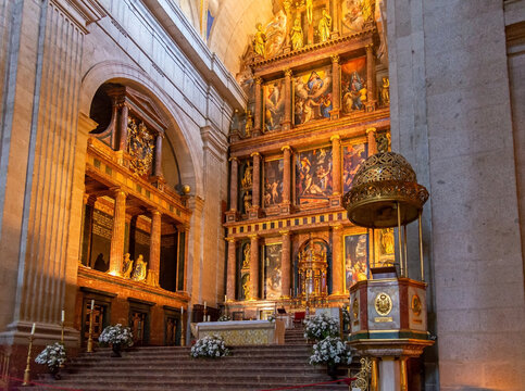 A centered view of the gilded high altar at the Basilica of San Lorenzo de El Escorial in Spain, featuring religious paintings, royal statues, and white flowers