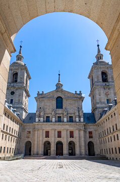 A sunny view of a symmetrical inner patio at El Escorial in Spain, featuring massive granite towers, uniform facades, and a grand basilica entrance. San Lorenzo