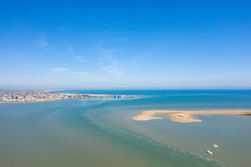 Aerial view of Baie de l'Orne near Ouistreham, showing sandy tidal flats, calm blue water, and distant town under a bright clear sky.