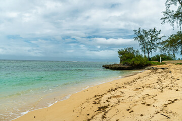 Secluded tropical beach with golden sand and turquoise ocean water under an overcast, cloudy sky near a rocky outcrop