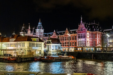 Obraz premium Railway station Amsterdam canal water boat Netherlands at night. Evening street houses architecture with lights in windows lanterns.