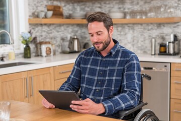Man in a wheelchair browses a tablet while sitting at a kitchen table with wooden cabinets