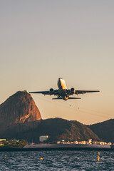 Passenger jet flying low over Guanabara Bay near Sugarloaf Mountain, Rio de Janeiro, Brazil, at golden hour. Dramatic aviation travel scene with copy space sky