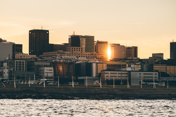 Golden hour city skyline behind airport fence and runway on the waterfront, small aircraft near terminal, sun reflection on towers, sea foreground, urban travel concept