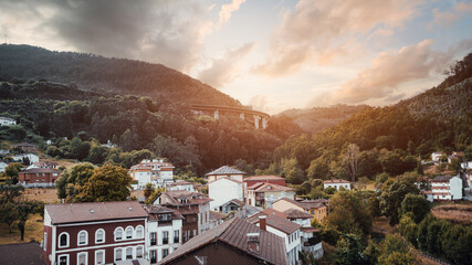 Sunset panorama of Salas, Asturias, along Camino de Santiago Primitivo: warm light on red roof village in green valley, forested mountains, clouds and curving viaduct