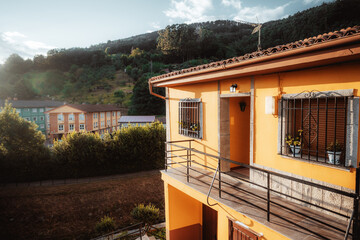 Sunlit orange countryside house with balcony, barred windows and tiled roof, overlooking green hillside and village buildings; wide blue sky copy space for text on the left