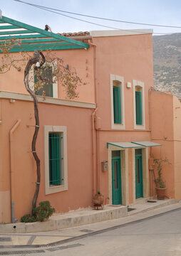 Renovated peach house with turquoise shutters in Archanes village. Minimalist Cretan architecture with a vine-covered pergola and mountain backdrop.