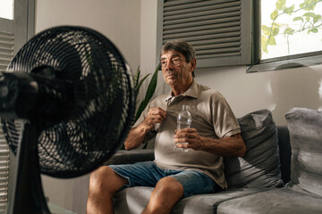 Senior man sits on a sofa at home using an electric fan to cool down on a hot day, holding a water...