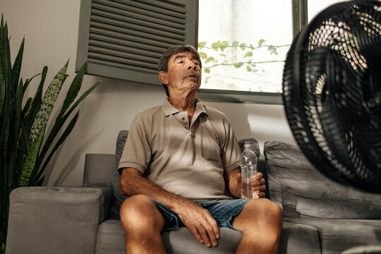 Senior man sits on a sofa at home using an electric fan to cool down on a hot day, holding a water bottle and resting, representing heat, hydration, comfort, and everyday wellbeing