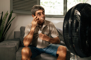 Senior man sits indoors feeling discomfort from heat while cooling himself with a fan, touching his head and resting, illustrating summer heat, fatigue, vulnerability, and everyday health concerns