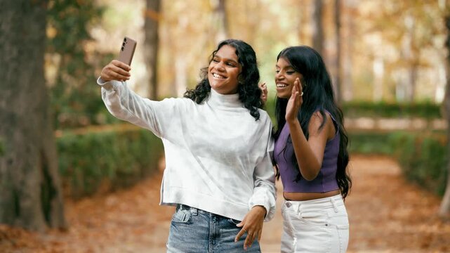 Two young indian friends smiling and waving while taking a selfie video with a smartphone. Happy women enjoying a beautiful autumn day outside