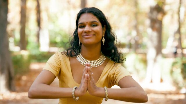 Graceful indian woman smiling and performing a traditional namaste greeting gesture. A dancer in a park expressing welcome and respect