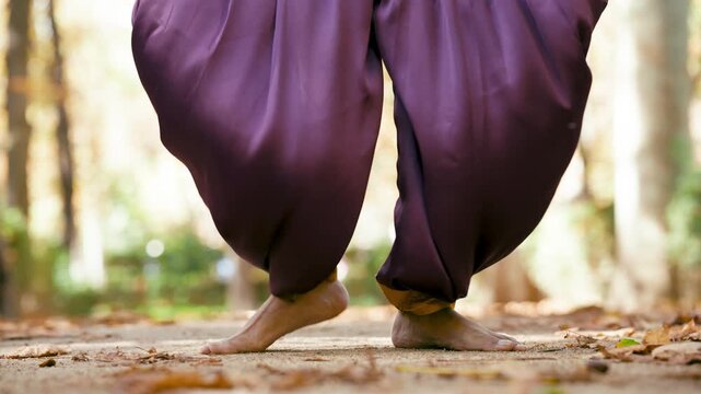 Close up of a barefoot dancer's feet performing traditional indian dance moves. The artist wears purple dhoti pants in a tranquil autumn park