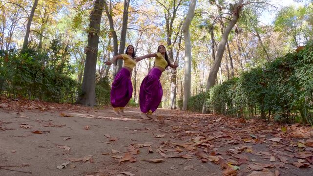 Two smiling friends in traditional indian attire performing a graceful and synchronized bollywood dance routine on a path in a beautiful autumn park