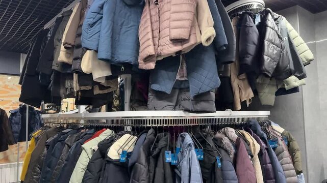 Rows Of Winter Coats Hanging, Shop Owner Sorting Preloved Inventory Beneath Crowded Rack, Mixed Textures Wool Denim Leather, Muted And Bright Colors, Cozy Cluttered Atmosphere, Sustainable.