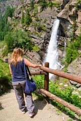 Woman hiking near Lillaz waterfalls in Cogne, Italy, admiring a powerful alpine cascade surrounded by rocky cliffs, green forest and wildflowers on a sunny summer day.