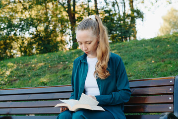 young beautiful redhead caucasian woman holds thick book in hands, reading book, sitting on bench in park, sunny summer day