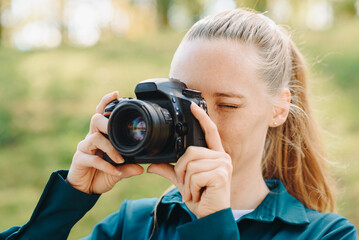 black dslr camera in hands, young pretty redhead caucasian woman photographer in green shirt at work outdoors in sunny summer day
