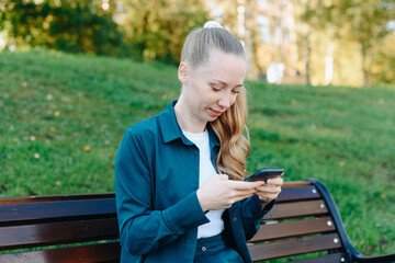young beautiful redhead caucasian woman holds smartphone in hands, texting, watching content, sitting on bench in park