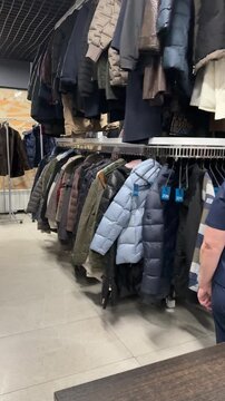 Rows Of Winter Coats Hanging, Shop Owner Sorting Preloved Inventory Beneath Crowded Rack, Mixed Textures Wool Denim Leather, Muted And Bright Colors, Cozy Cluttered Atmosphere, Sustainable.